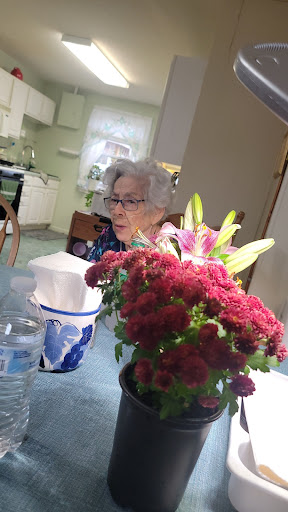 An elderly woman seated at a table surrounded by vibrant flowers in a cozy kitchen setting.