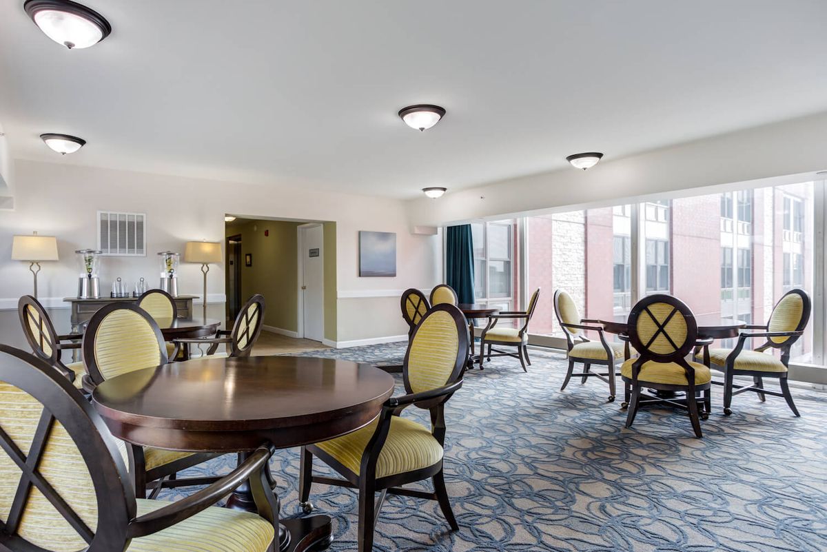 A well-lit room with round tables and cushioned chairs, featuring large windows and patterned carpet.
