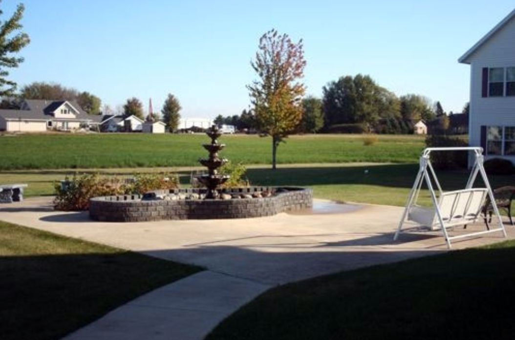 A scenic outdoor area with a fountain, surrounded by a pathway and lush fields.