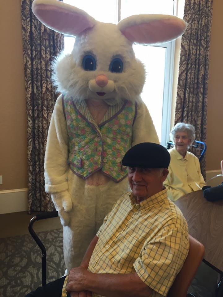 An Easter Bunny in costume stands next to an elderly man seated at a table during a festive indoor event.