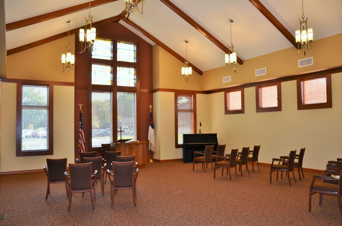 An interior view of a chapel with chairs arranged around a piano and large windows allowing natural light.