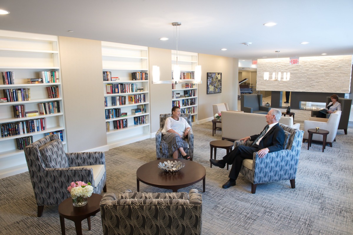 Two people having a conversation in a library setting with bookshelves and comfortable seating.
