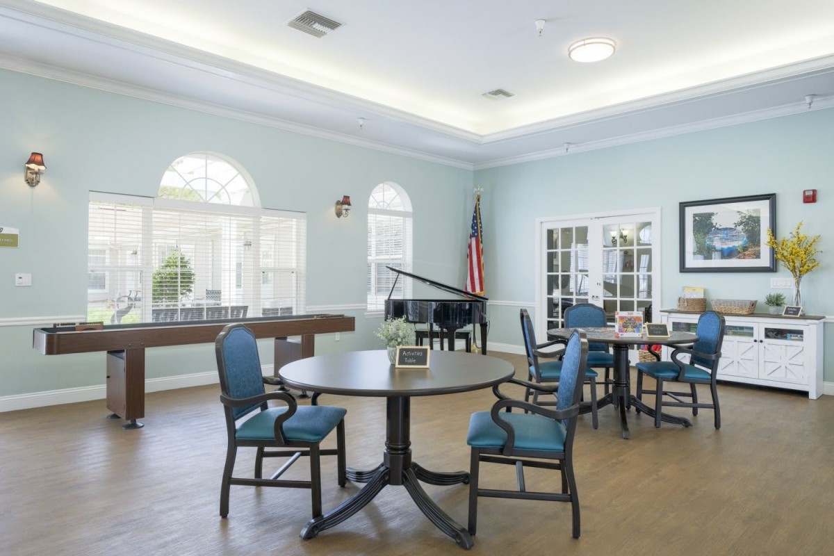 A bright recreation room featuring a piano, tables, and chairs.