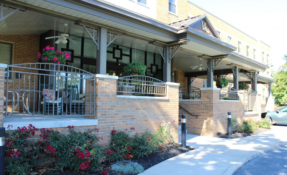 The image shows the entrance of a building with a porch and brick walls, surrounded by landscaping and flowers.