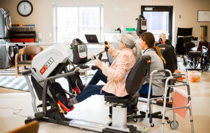 An elderly person engaging in a workout session using specialized exercise equipment with assistance from a caregiver.
