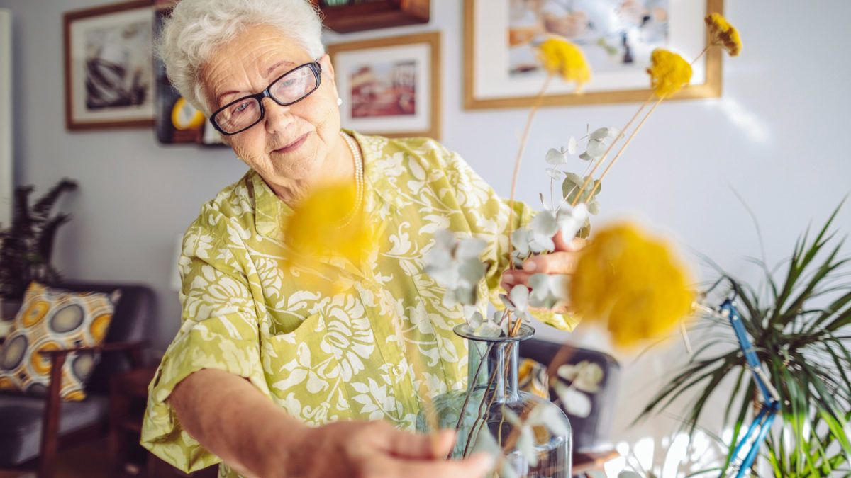 An elderly woman wearing glasses arranges a bouquet of yellow flowers at home.