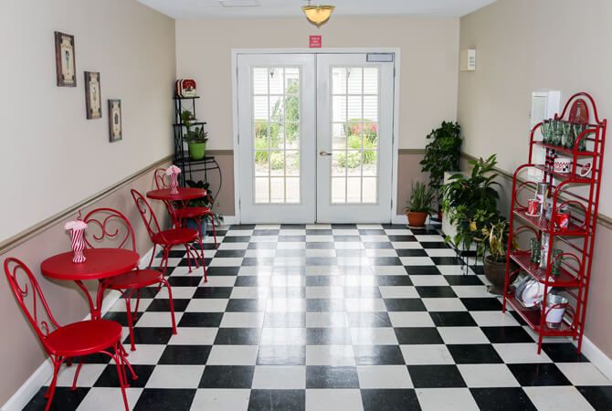 A hallway with black and white checkered flooring, featuring red chairs and green plants along its sides.