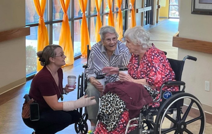Three women engage in a warm conversation, two seated in wheelchairs and one kneeling beside them, sharing a moment of companionship indoors.