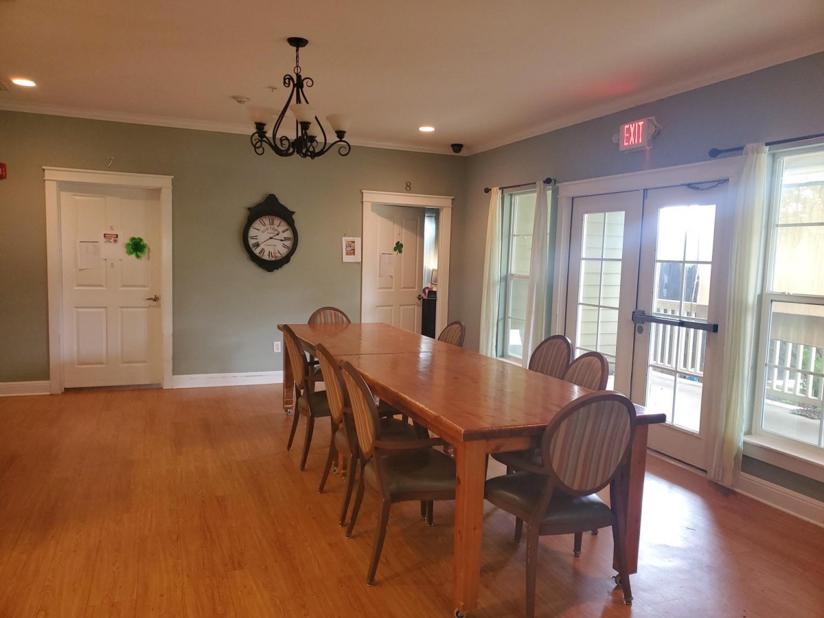 A cozy dining room with a long wooden table and elegant chairs, featuring a decorative chandelier and wall clock.