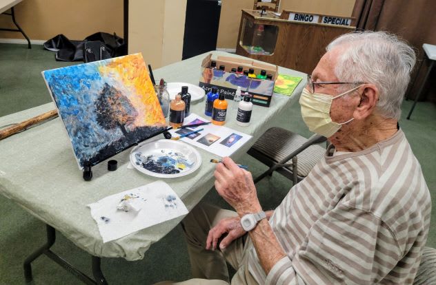 An elderly person wearing a mask is engaged in painting a colorful canvas indoors, surrounded by art supplies.