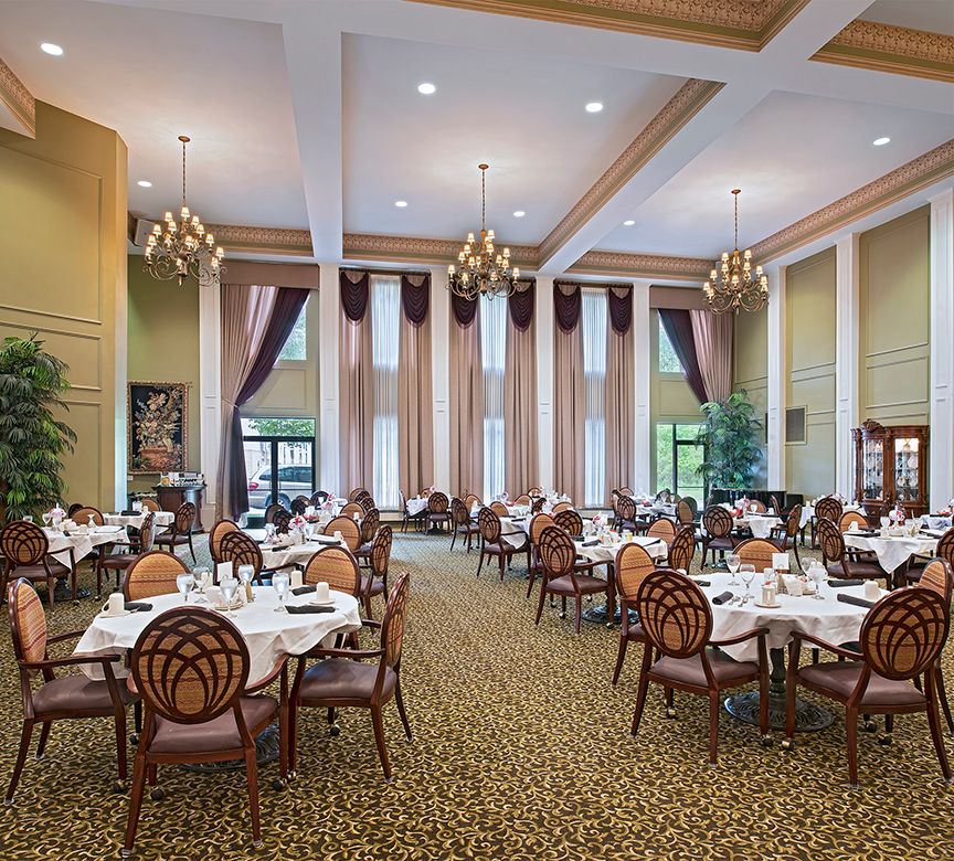 A spacious and elegantly decorated dining room with multiple tables and chairs, featuring tall windows and chandeliers.