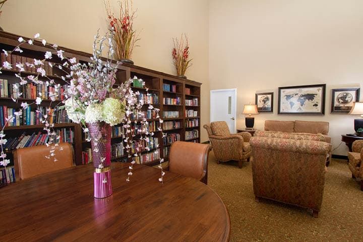 A cozy library room with bookshelves, comfortable chairs, and a table adorned with decorative flowers.