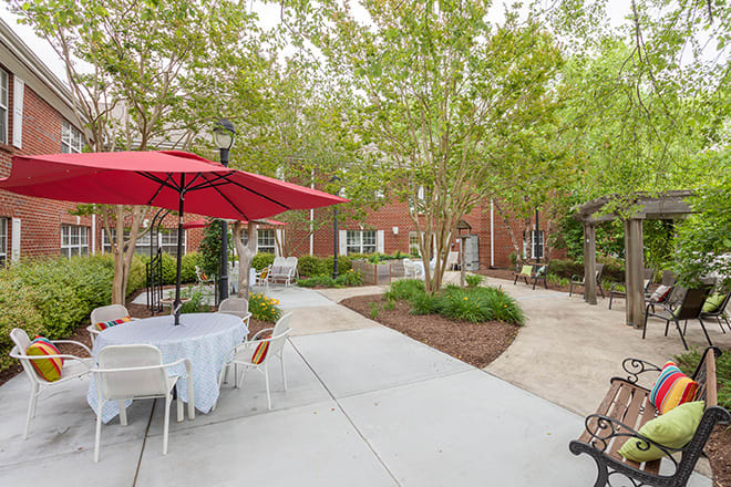 A peaceful outdoor patio with tables and chairs, featuring red umbrellas and surrounded by greenery.