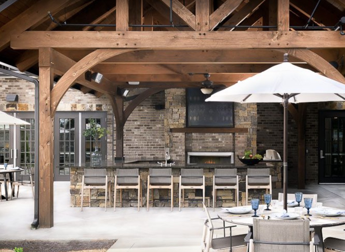 A stylish outdoor kitchen and patio area featuring stone walls, wooden beams, bar seating, and an umbrella.