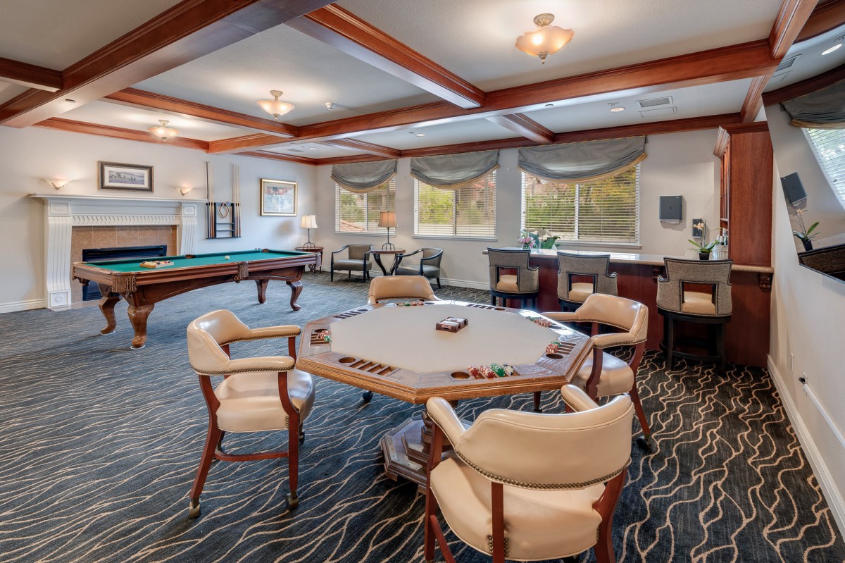 A spacious game room featuring a poker table and a pool table, with large windows and comfortable seating.