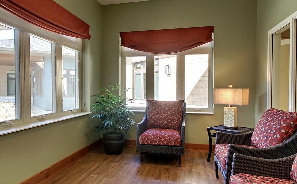 A cozy corner of a living room featuring a patterned chair, a side table with a lamp, and large windows with red curtains.