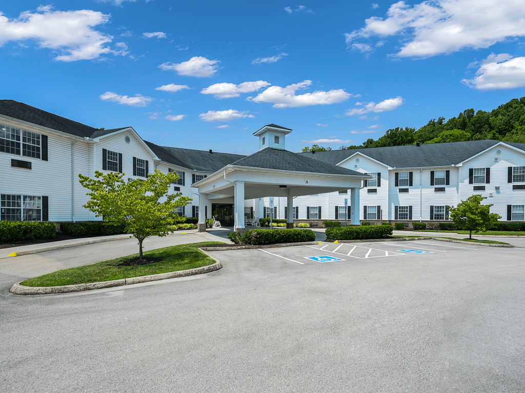 A large building with a covered entrance, surrounded by a parking lot, trees, and a clear blue sky with clouds.
