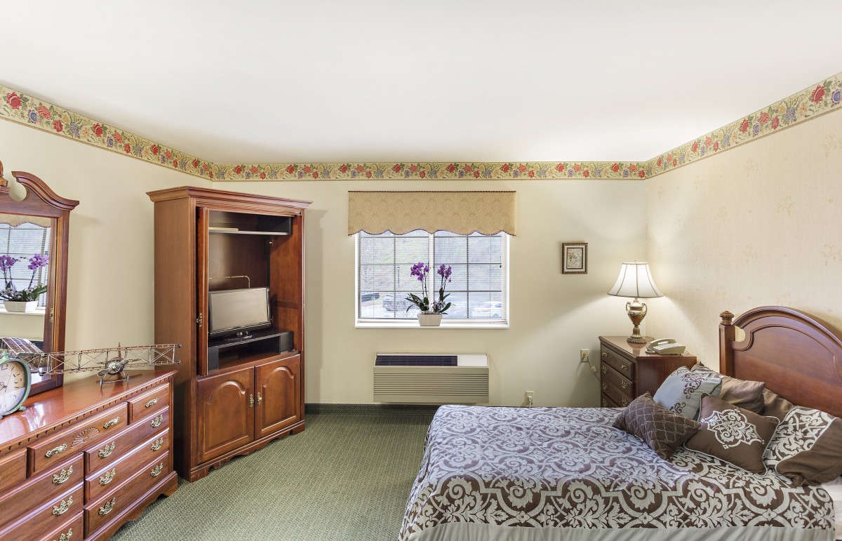 A cozy bedroom with a wooden cabinet, a TV, a large window, and a bed decorated with patterned bedding.