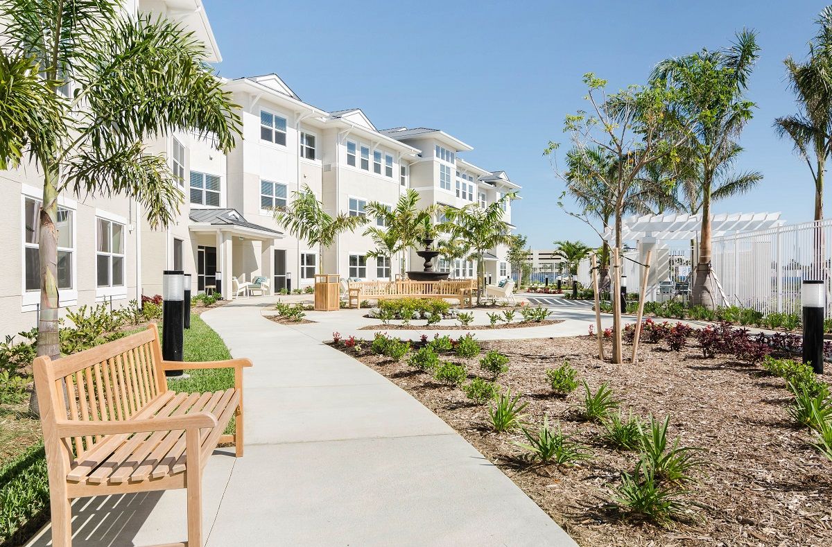 A modern apartment complex with well-maintained landscaping and palm trees along a walkway, featuring benches for seating.