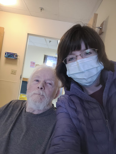 A selfie taken in a hospital setting, featuring an older adult and a caregiver, both wearing face masks.