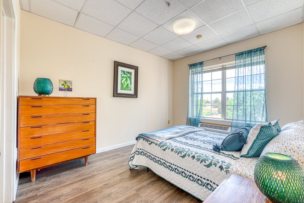 A cozy bedroom with a bed, wooden dresser, and a large window with blue curtains.