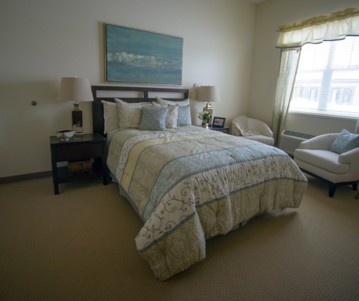 A cozy bedroom with a neatly made bed, bedside tables with lamps, artwork above the bed, and a comfortable chair by the window.
