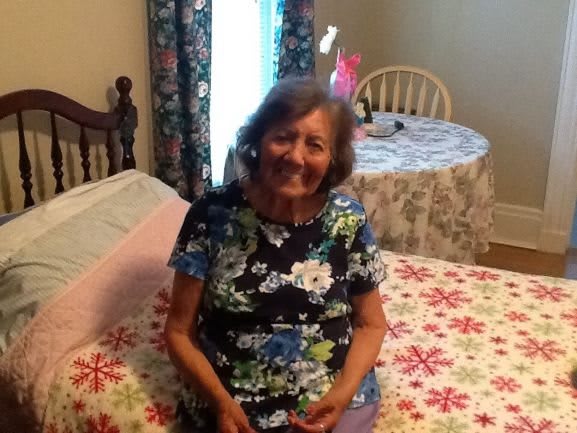 An elderly woman sitting on a bed, smiling in a cozy room.
