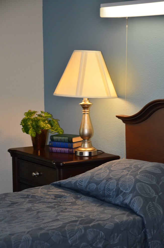A cozy bedroom setup with a lamp on a nightstand next to books and a plant.