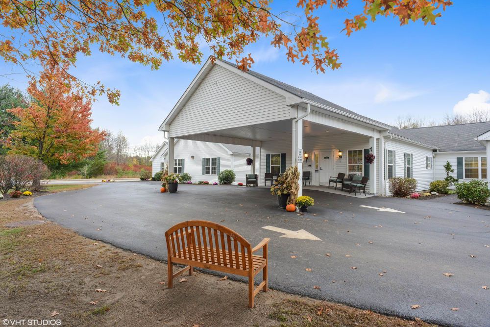 A white building with a covered entrance, surrounded by autumn trees and a wooden bench in the foreground.