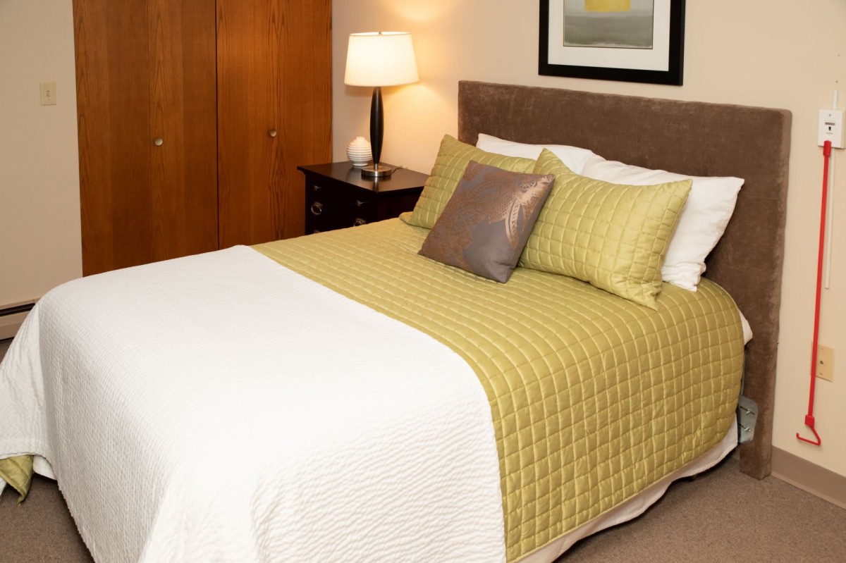 A cozy bedroom with a neatly made bed, featuring green and white bedding, accompanied by a nightstand and lamp.