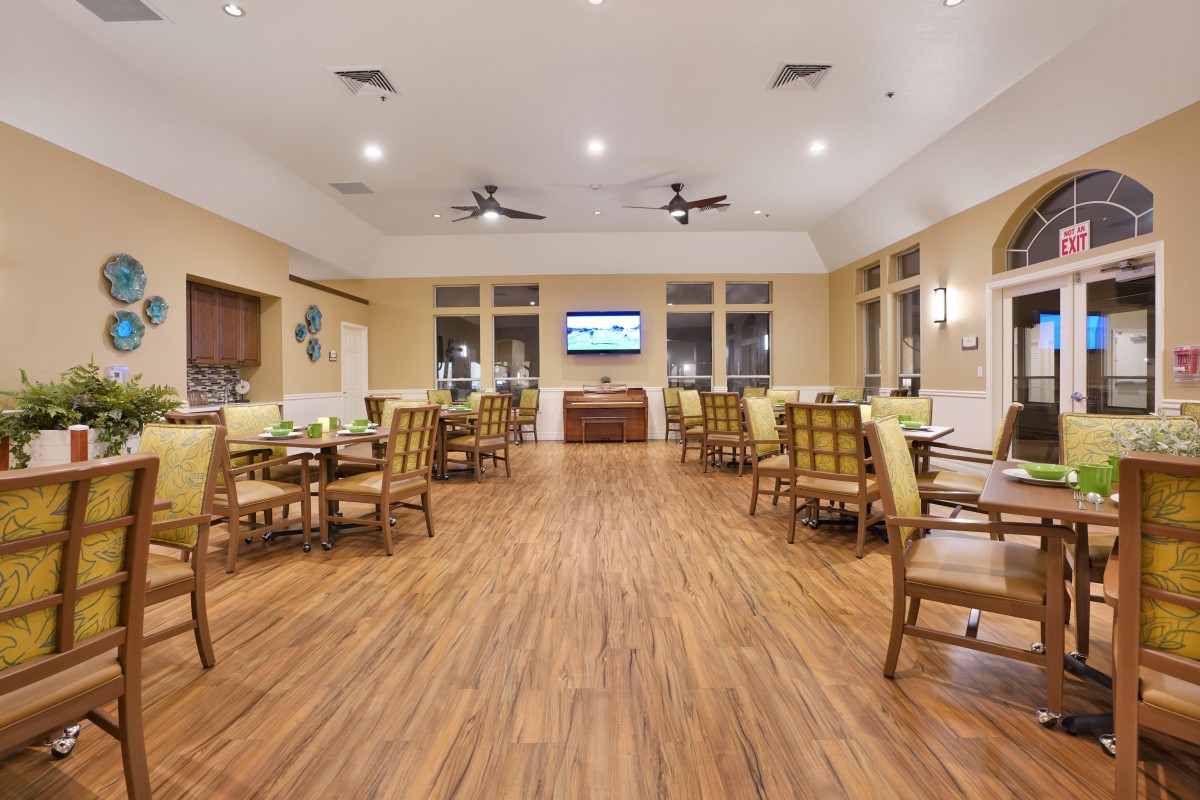 A spacious dining area with wooden flooring, tables, and chairs, featuring ceiling fans and a television on the wall.