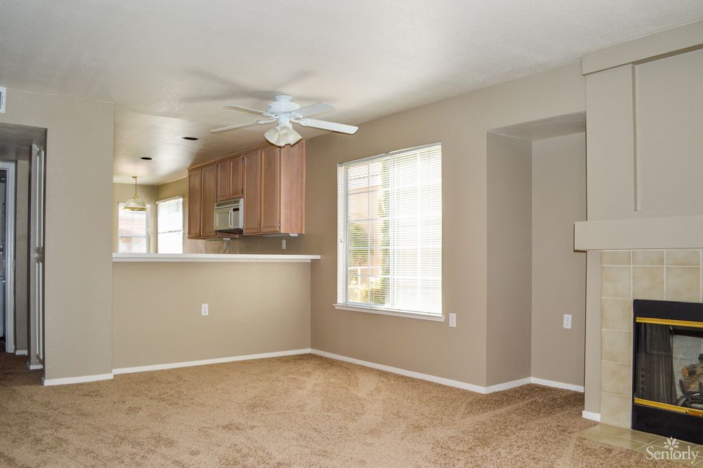An empty living room with carpet flooring, a large window, and a view into the kitchen area.