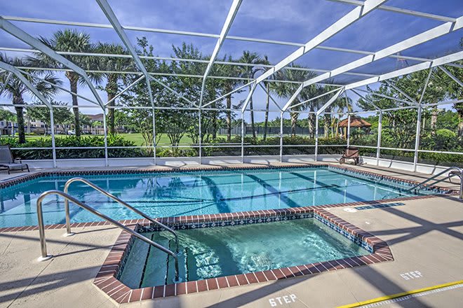 A screened outdoor area featuring a swimming pool and a hot tub surrounded by trees and blue skies.