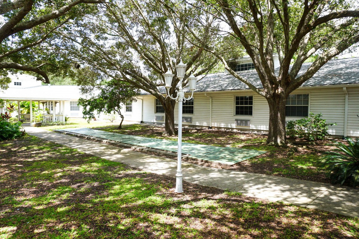 A peaceful outdoor scene featuring a house surrounded by trees, with a lamp post and benches along a pathway.