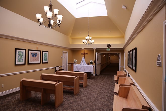 A serene chapel interior with wooden pews, a cross at the altar, and soft lighting from chandeliers and a skylight.