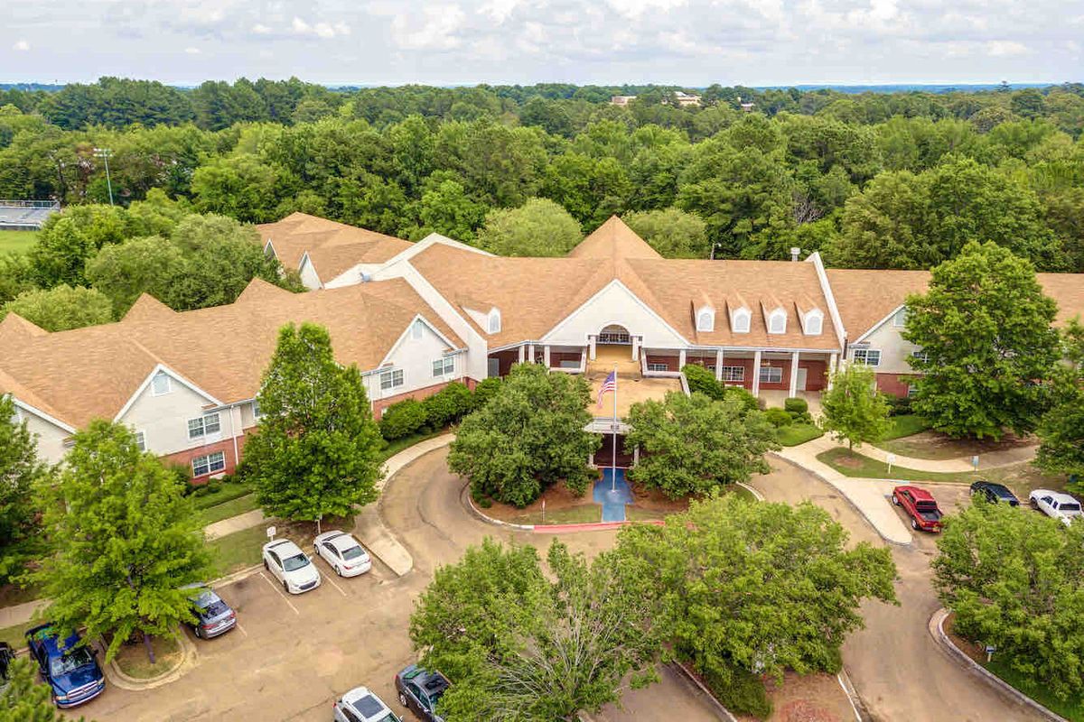 Aerial view of a large building surrounded by trees with cars parked along the driveway.
