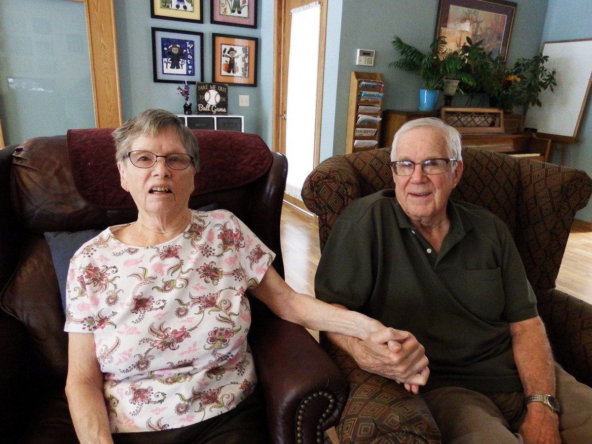 A senior couple sitting in a cozy living room, holding hands and surrounded by family photos.