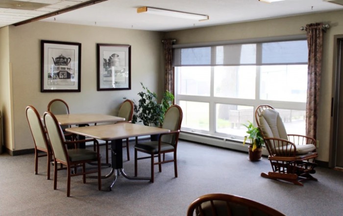 A cozy room with tables and chairs, featuring large windows and plants.
