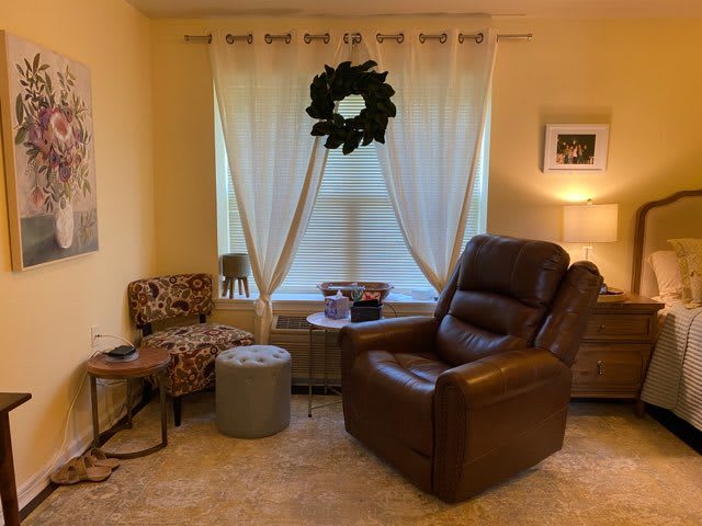 A cozy living room featuring a leather recliner and patterned chair, accented by light curtains and a decorative wreath.