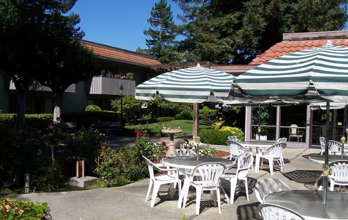 A sunny outdoor patio area with tables, chairs, and striped umbrellas surrounded by a garden.