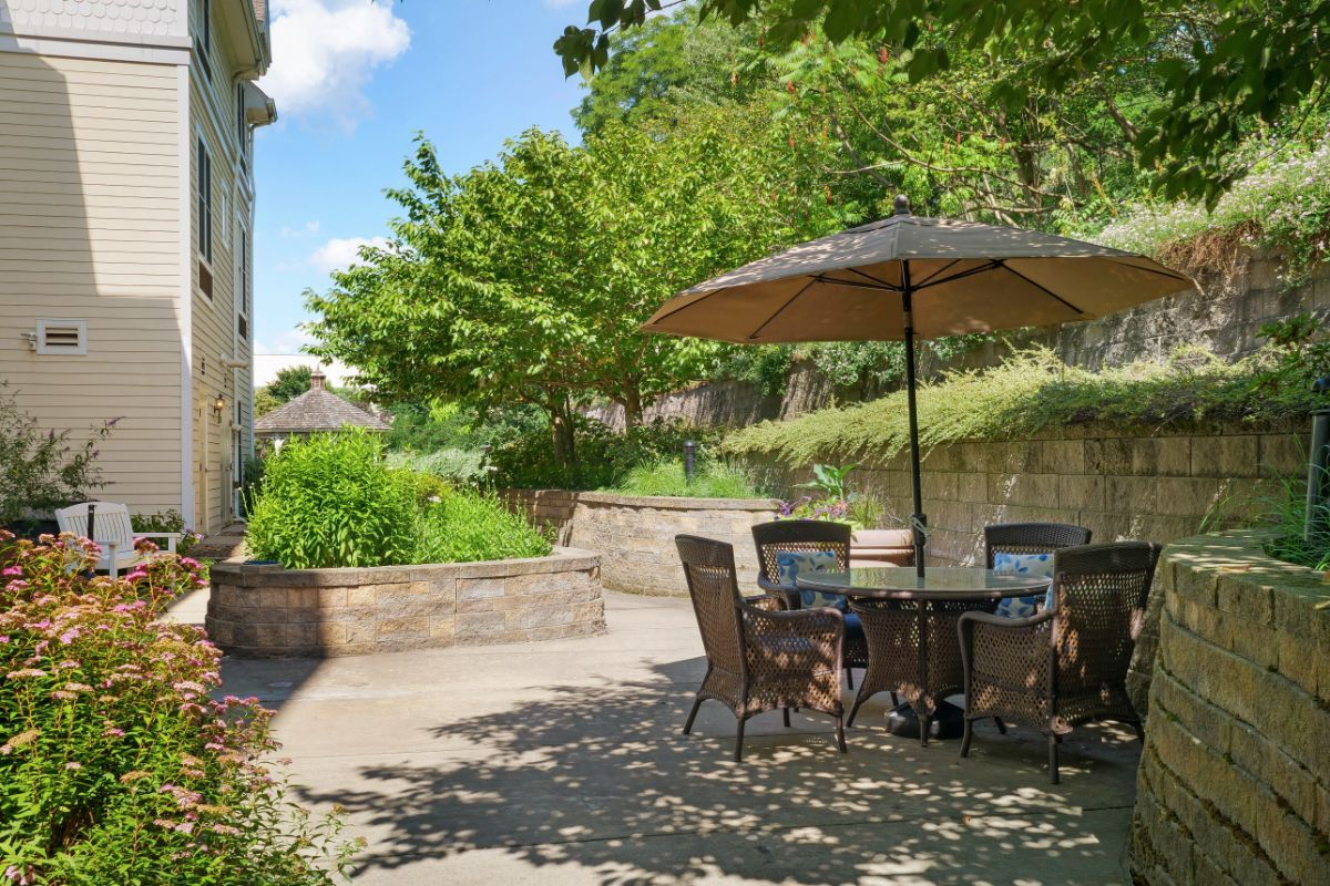 A peaceful patio with outdoor furniture and a large umbrella, surrounded by lush greenery on a sunny day.