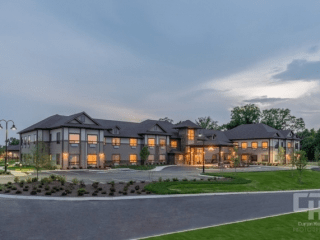 A luxurious two-story mansion illuminated in the evening, surrounded by landscaped grounds.