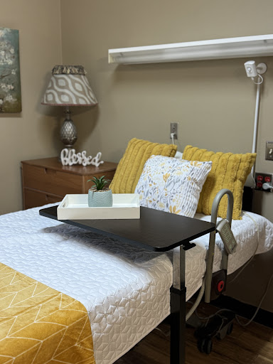 A cozy hospital room featuring a neatly made bed with yellow and white bedding, accompanied by a bedside lamp and a table tray.