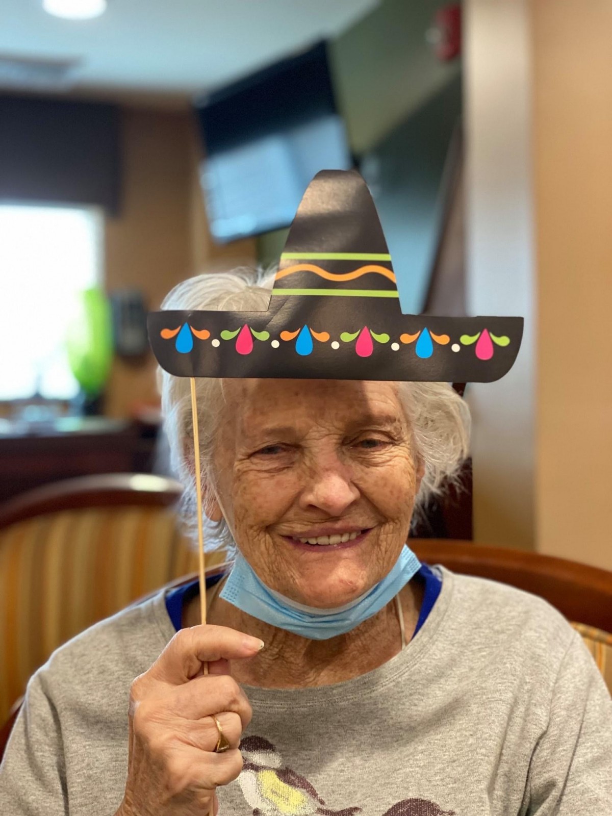 An elderly woman smiling while holding a decorative hat in an indoor setting.