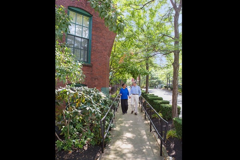 Two people walk along a tree-lined sidewalk beside a brick building on a sunny day.