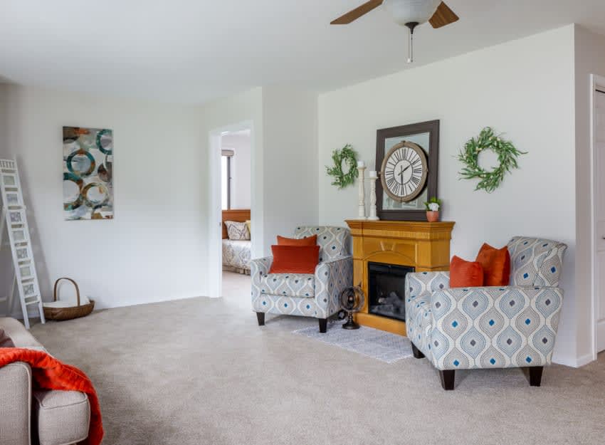 A cozy living room featuring patterned armchairs, a wooden fireplace with a large clock above it, and decorative wreaths on the wall.