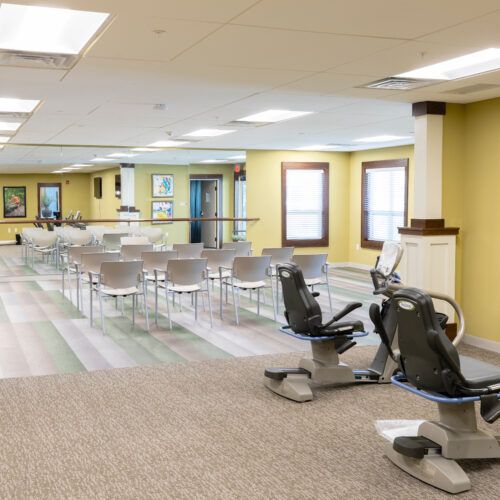A spacious gym interior featuring exercise equipment, chairs, mirrors, and windows.