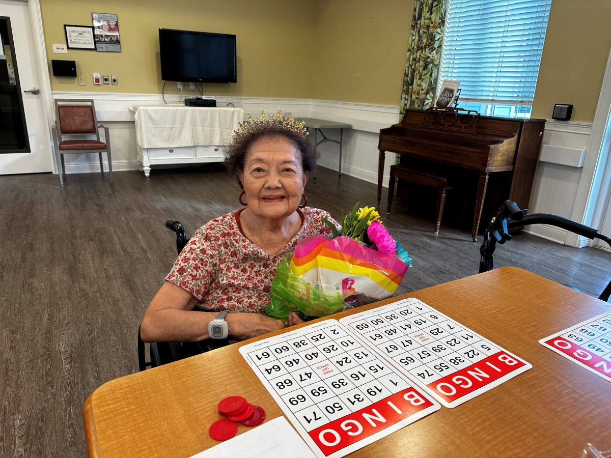An elderly woman in a wheelchair holding flowers and playing bingo indoors.
