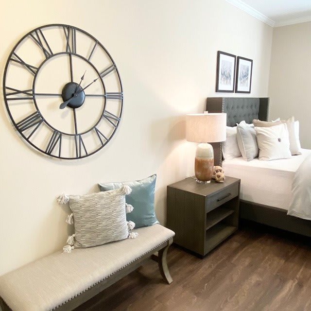 A cozy bedroom with a large wall clock, comfortable bench, and neatly arranged pillows on the bed.