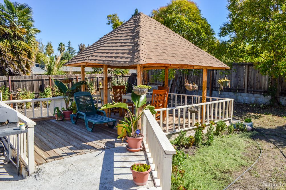 A sunny outdoor garden space featuring a wooden gazebo, surrounded by plants and patio furniture.